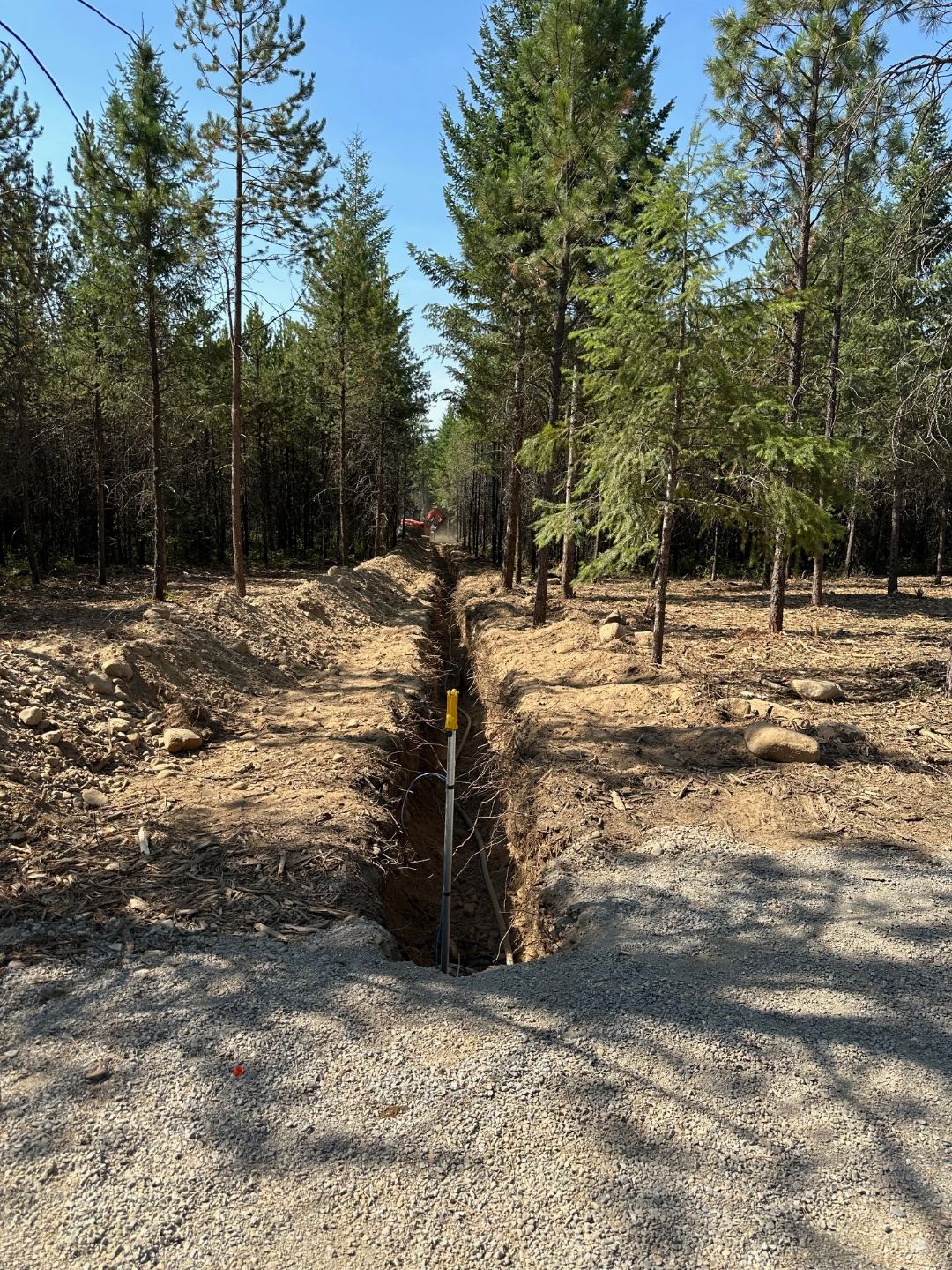 Deep utility trench cut through a forested lot with riser pipe in foreground and excavator at the end.