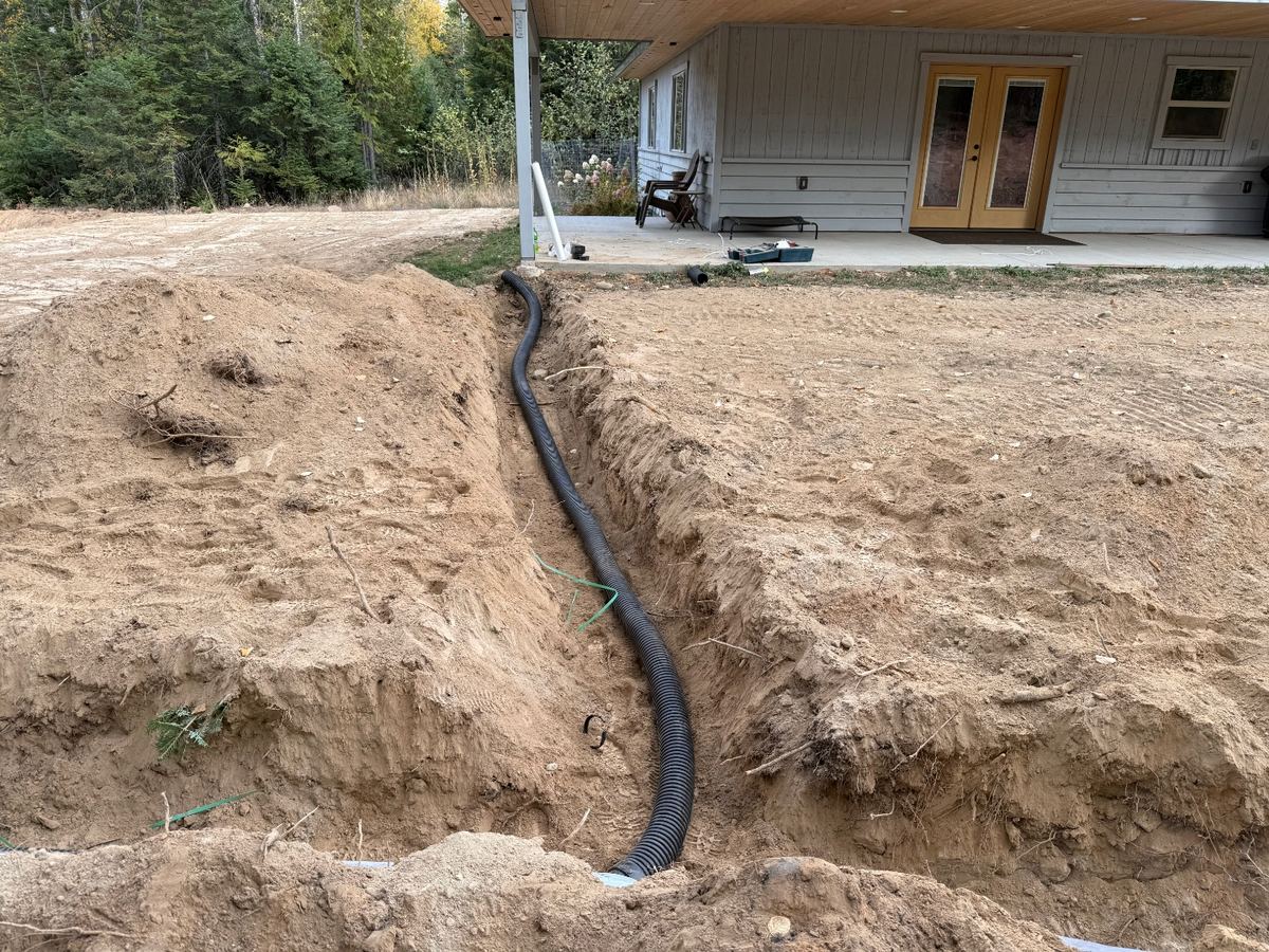 Black corrugated foundation drain line running through a freshly cut drainage swale from a house, graded to daylight.
