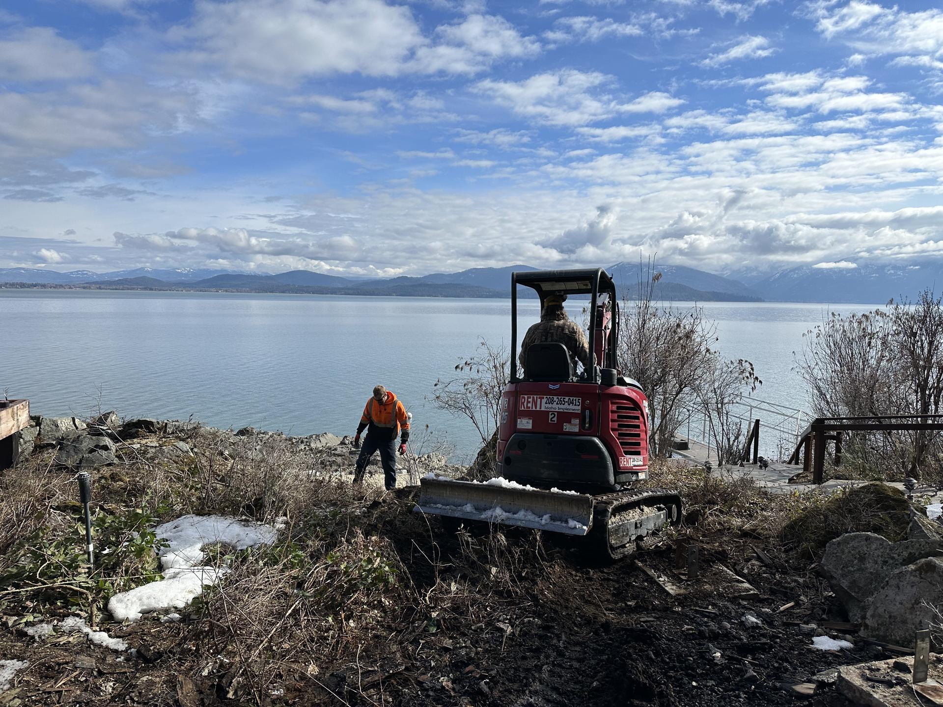 Mini excavator on lakefront building boulder retaining wall Pend Oreille Lake backdrop