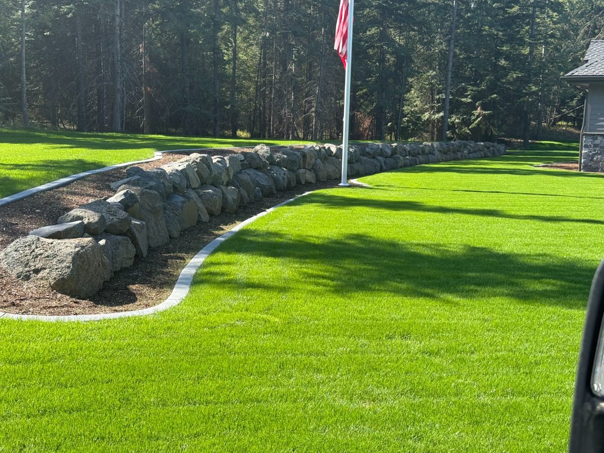 Curved granite boulder retaining wall with cut-stone mow curb, finished lawn and flagpole — Premier Land Service retaining walls, North Idaho
