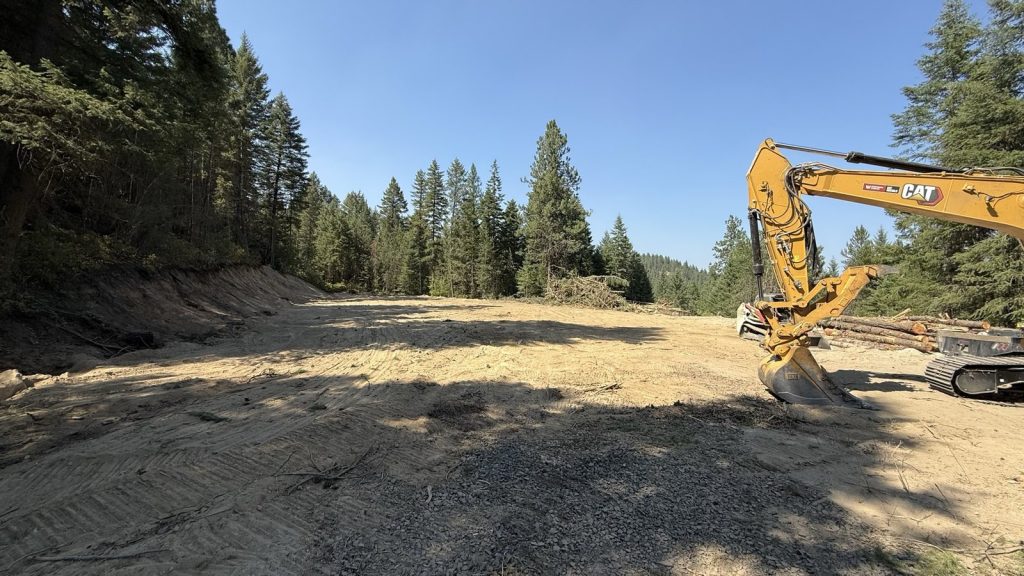 Cleared and graded house pad with stacked logs and forested backdrop, Sagle Idaho