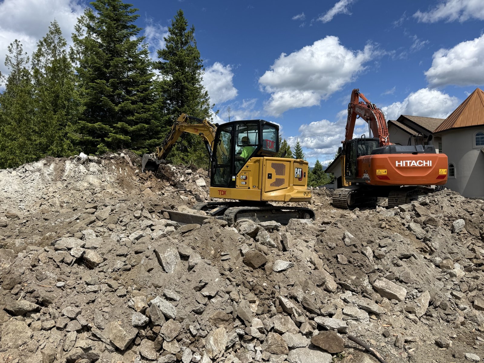 Mini excavator and full-size excavator breaking rock and grading a house pad above Sandpoint, Idaho
